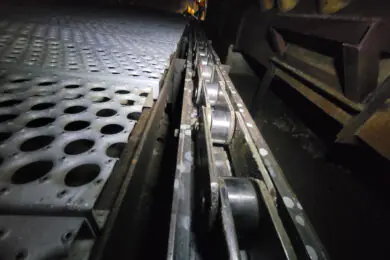 Close-up of an e-coat oven conveyor chain and roller track next to perforated floor grates.